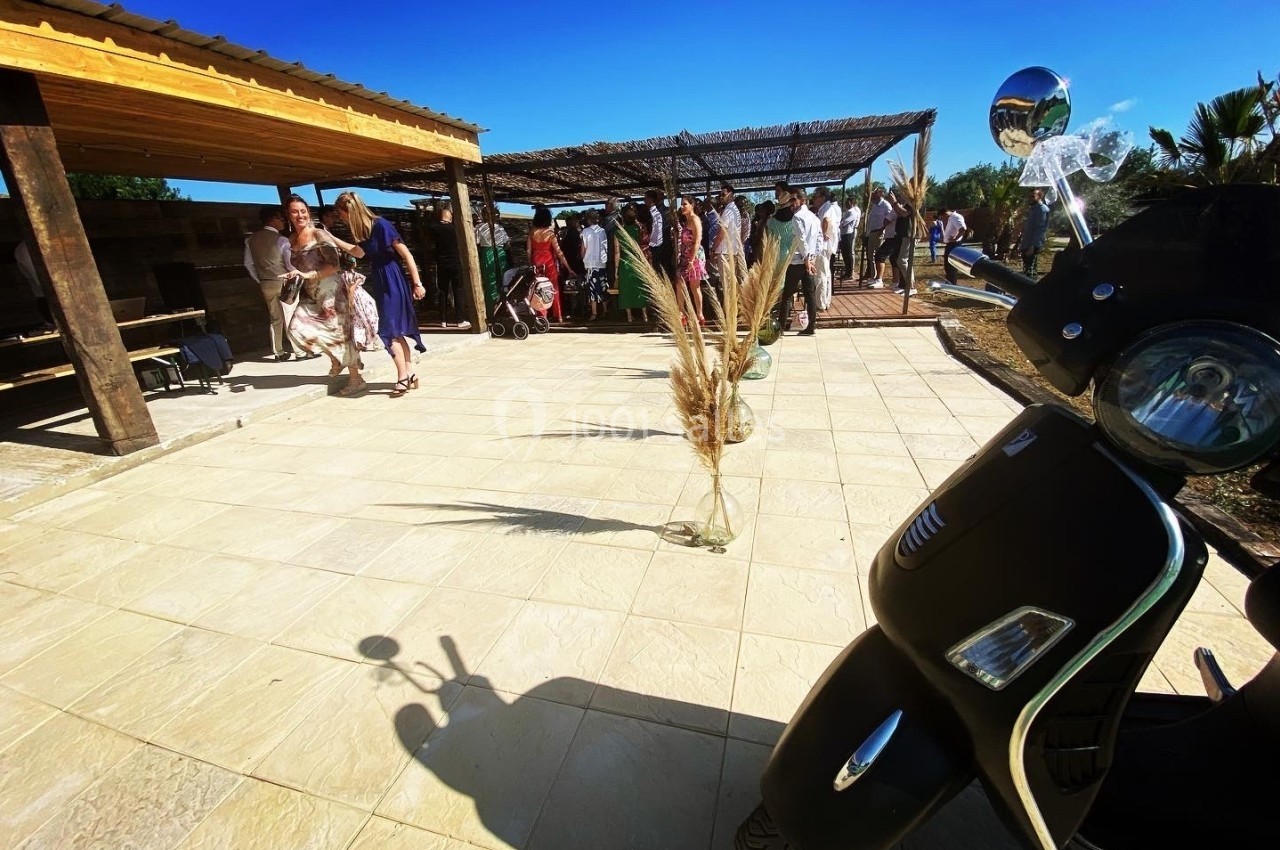 Un groupe de personnes rassemblées sous une pergola en bois, avec un scooter noir au premier plan sur une terrasse…