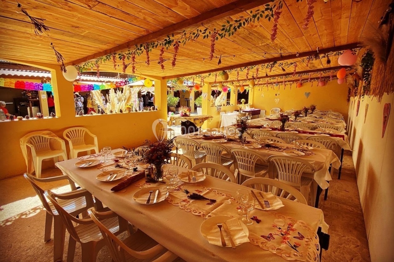 Salle décorée pour un repas festif, avec longues tables dressées, chaises blanches et guirlandes suspendues sous un toit en…