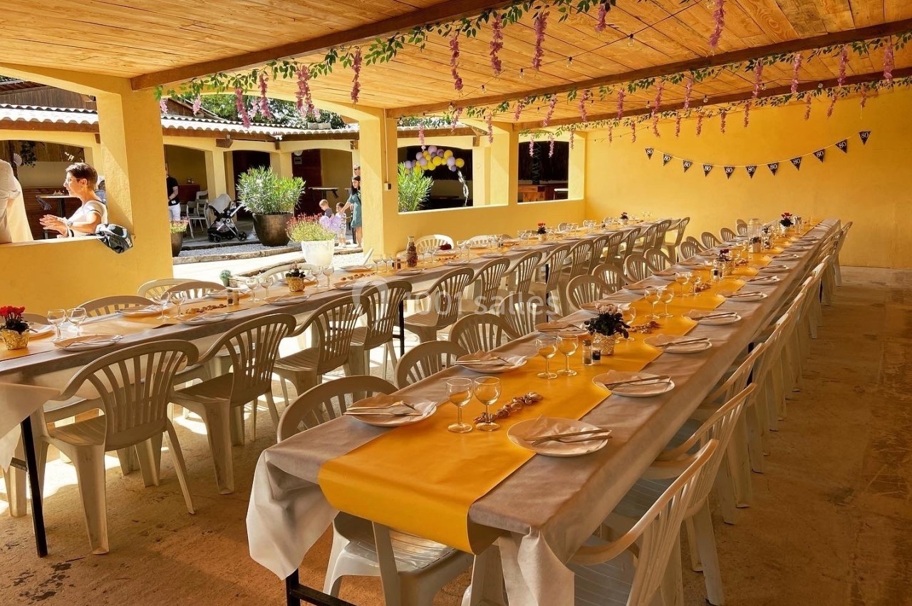 Tables longues dressées avec nappes jaunes et blanches sous un auvent décoré de guirlandes fleuries.