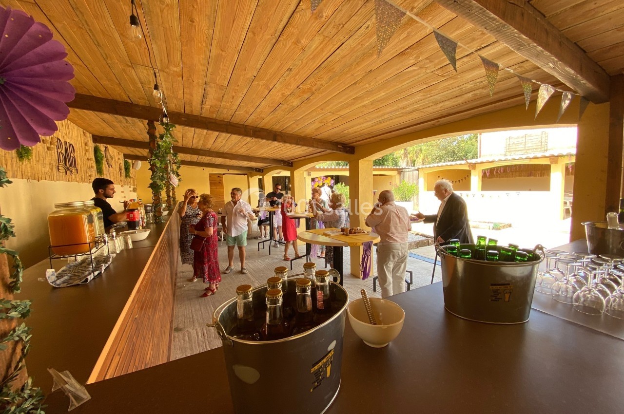 Des personnes discutent autour de tables dans un espace couvert en bois, avec un bar et des boissons en libre-service.