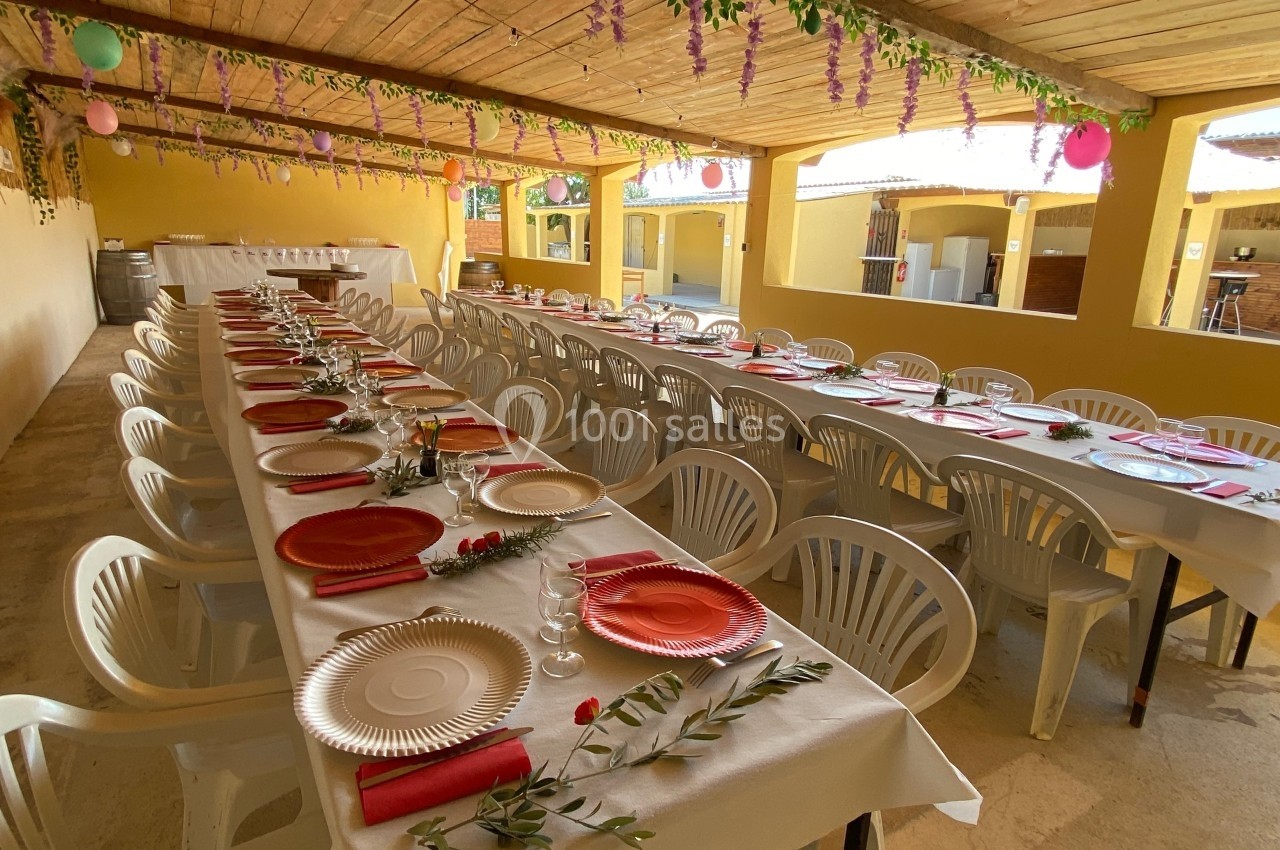 Salle décorée pour un repas festif avec de longues tables alignées, nappes blanches et assiettes colorées.