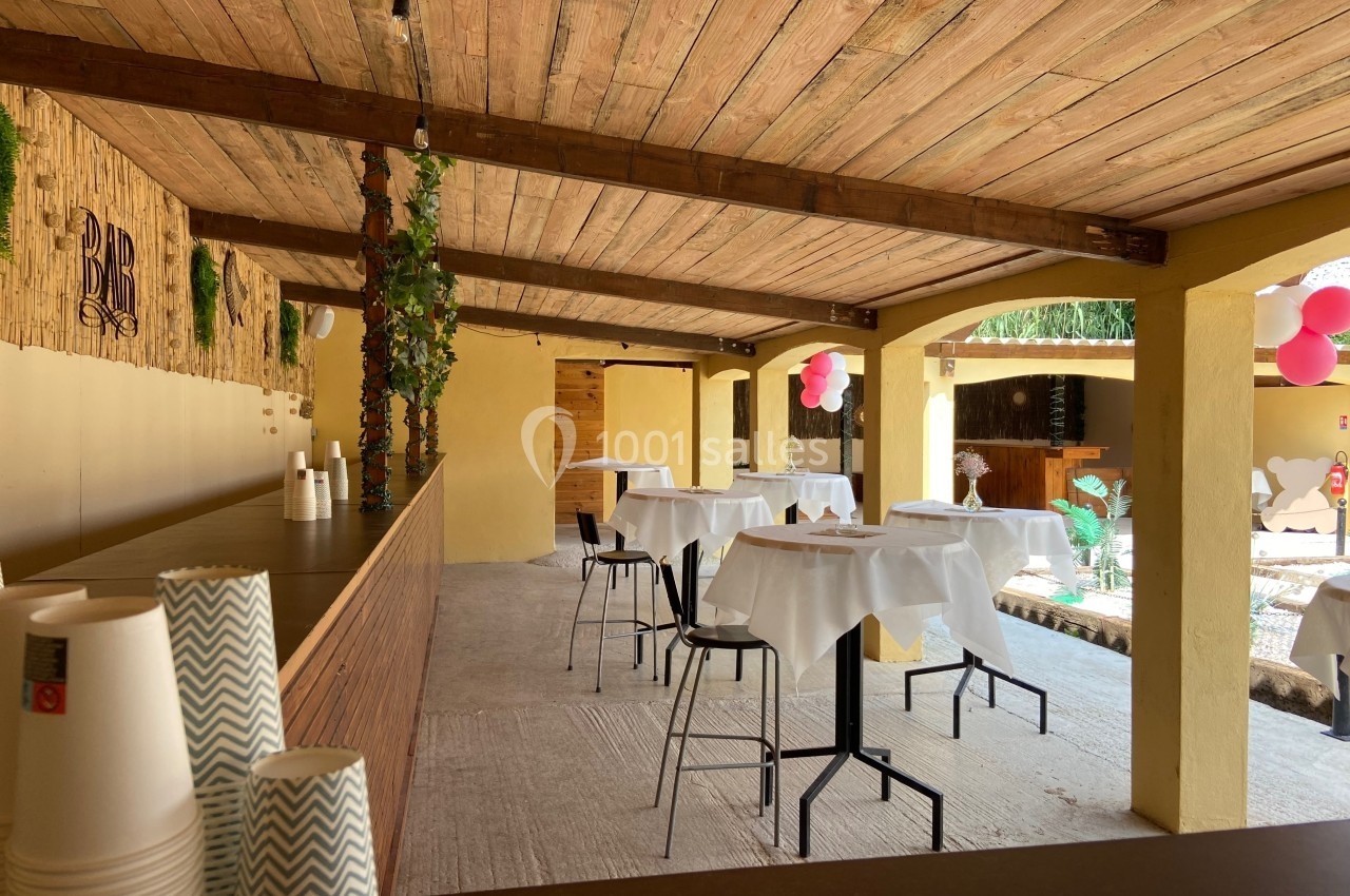 Terrasse couverte avec tables hautes dressées, bar en bois, décorations végétales et ballons roses en arrière-plan.