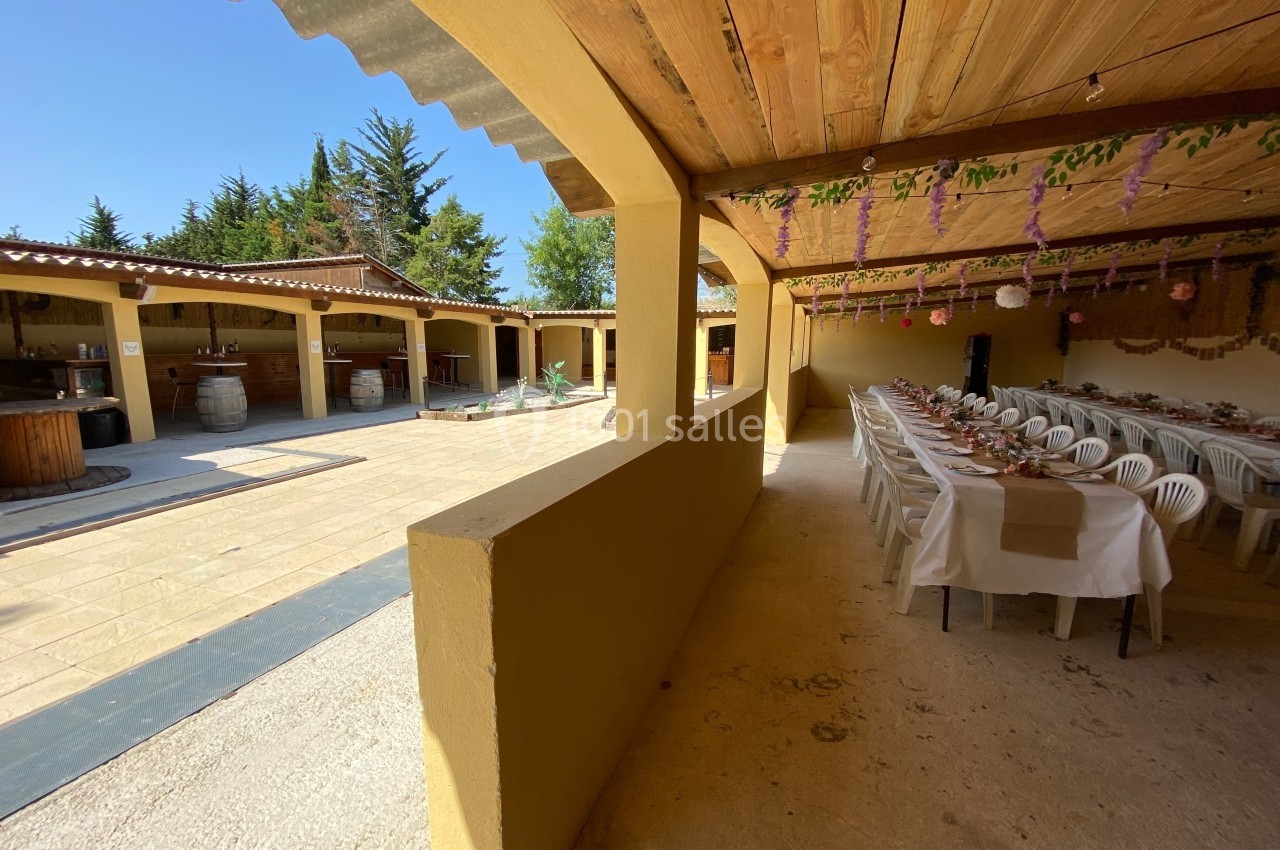 Salle extérieure couverte avec tables dressées pour un repas, donnant sur une cour avec bar et décorations suspendues.