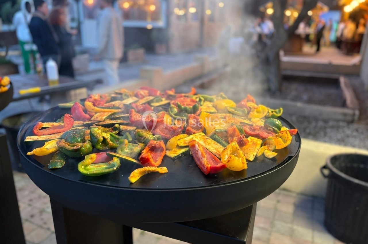 Légumes colorés grillant sur une plancha en extérieur, avec des lumières et des personnes en arrière-plan flou.