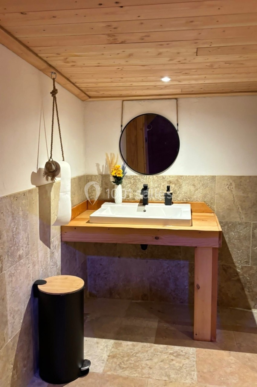 Lavabo en bois avec miroir rond, vase décoratif et accessoires, dans une salle de bain aux murs en pierre et plafond en bois.