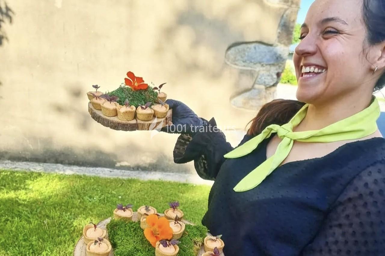 Une femme souriante présente des amuse-bouches décorés de fleurs sur des plateaux en bois, en extérieur.