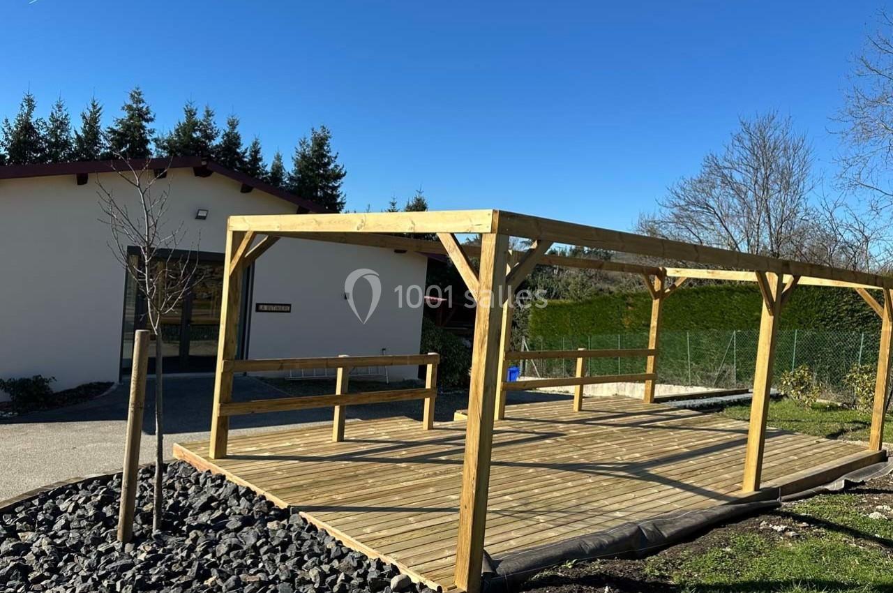 Structure en bois rectangulaire sur une terrasse ensoleillée, entourée de végétation et d'un bâtiment blanc en arrière-plan.