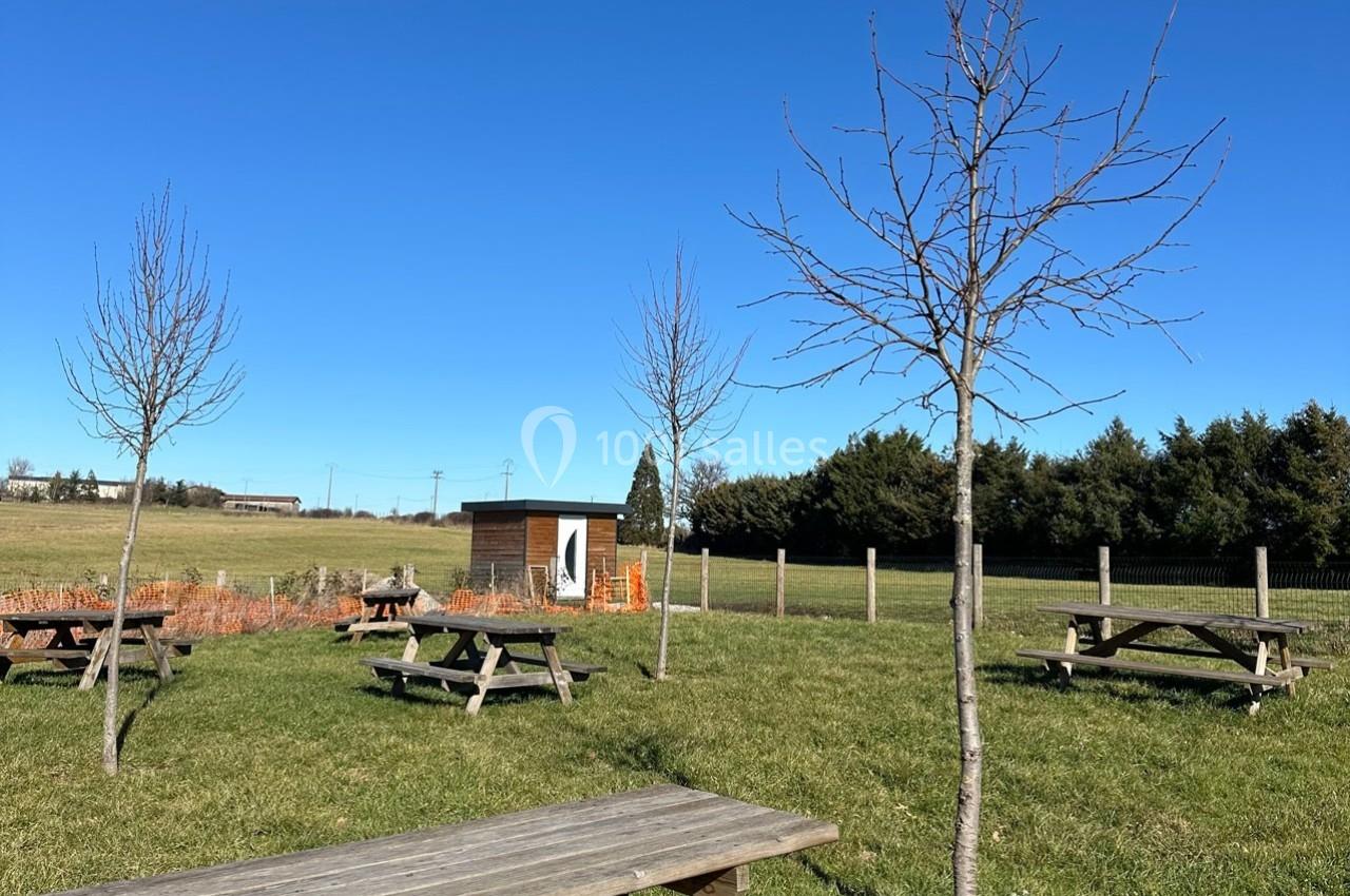 Tables de pique-nique en bois disposées sur une pelouse avec des arbres, sous un ciel bleu dégagé.