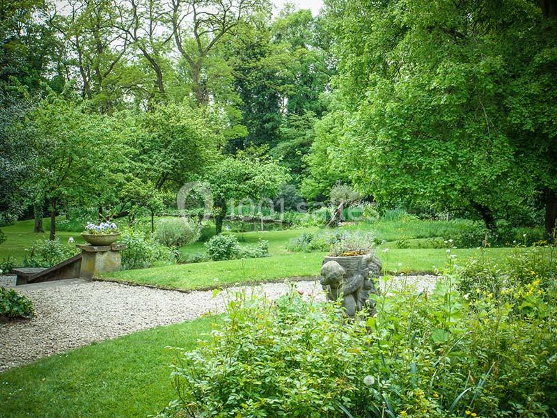 Jardin verdoyant avec pelouse, parterres fleuris, arbres et statue décorative en pierre au premier plan.