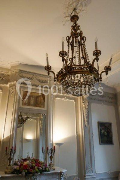 Lustre en métal doré et cristaux suspendu dans une pièce au décor classique avec moulures et cheminée ornée de fleurs.