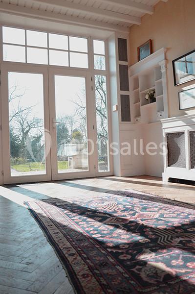 Salon lumineux avec parquet, tapis coloré, cheminée blanche et grande baie vitrée donnant sur un jardin.