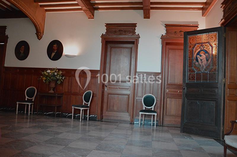 Salle avec parquet et murs en bois, trois portes, chaises alignées et une porte vitrée entrouverte ornée d'un motif.