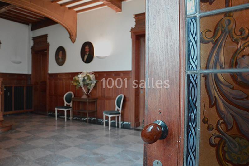 Vue d'une entrée avec porte en bois ouverte, sol en marbre, deux chaises et portraits accrochés sur des murs lambrissés.