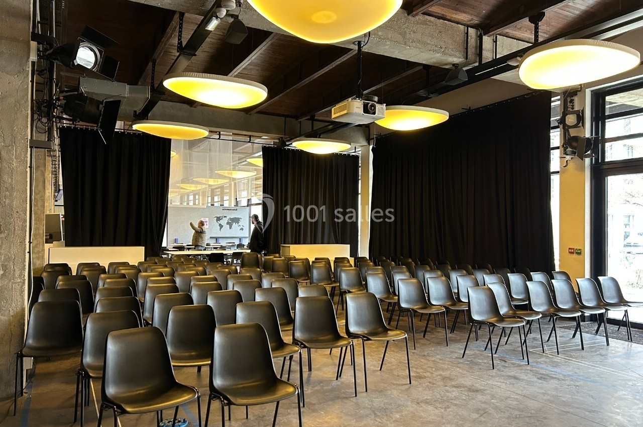 Salle de conférence avec rangées de chaises noires, éclairage suspendu et rideaux noirs en arrière-plan.