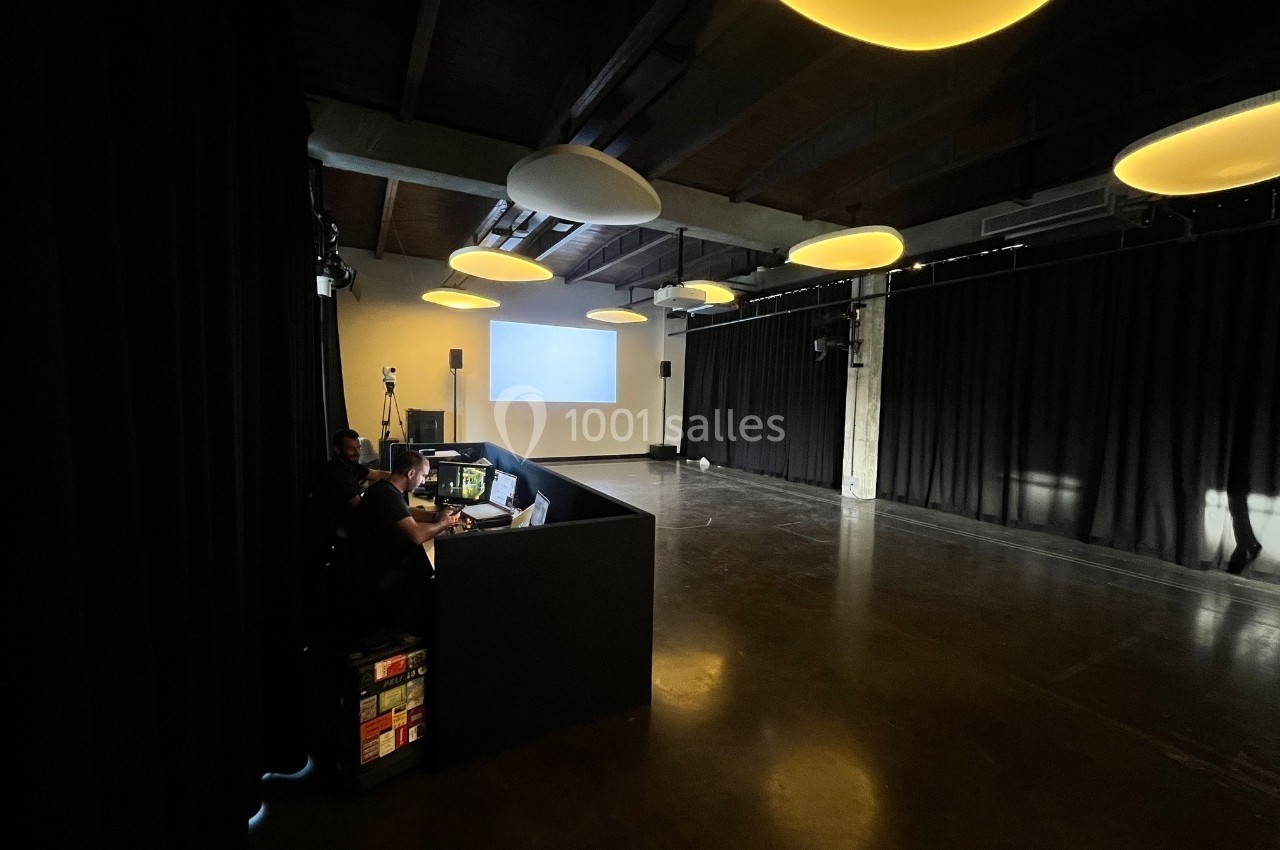 Salle sombre avec un espace de projection, un écran blanc, des rideaux noirs et une régie technique éclairée.