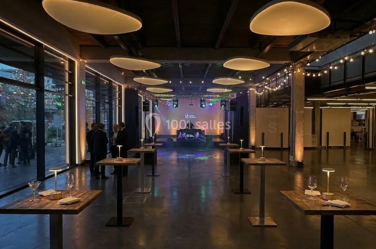 Salle événementielle moderne avec tables hautes en bois, éclairage tamisé et scène au fond.