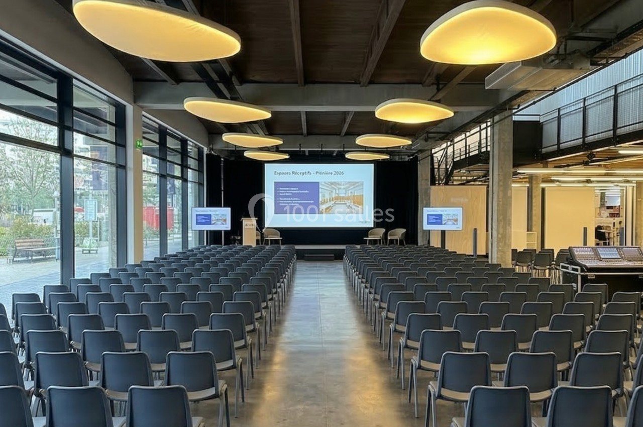 Salle de conférence moderne avec rangées de chaises vides, écran de projection et éclairage suspendu.