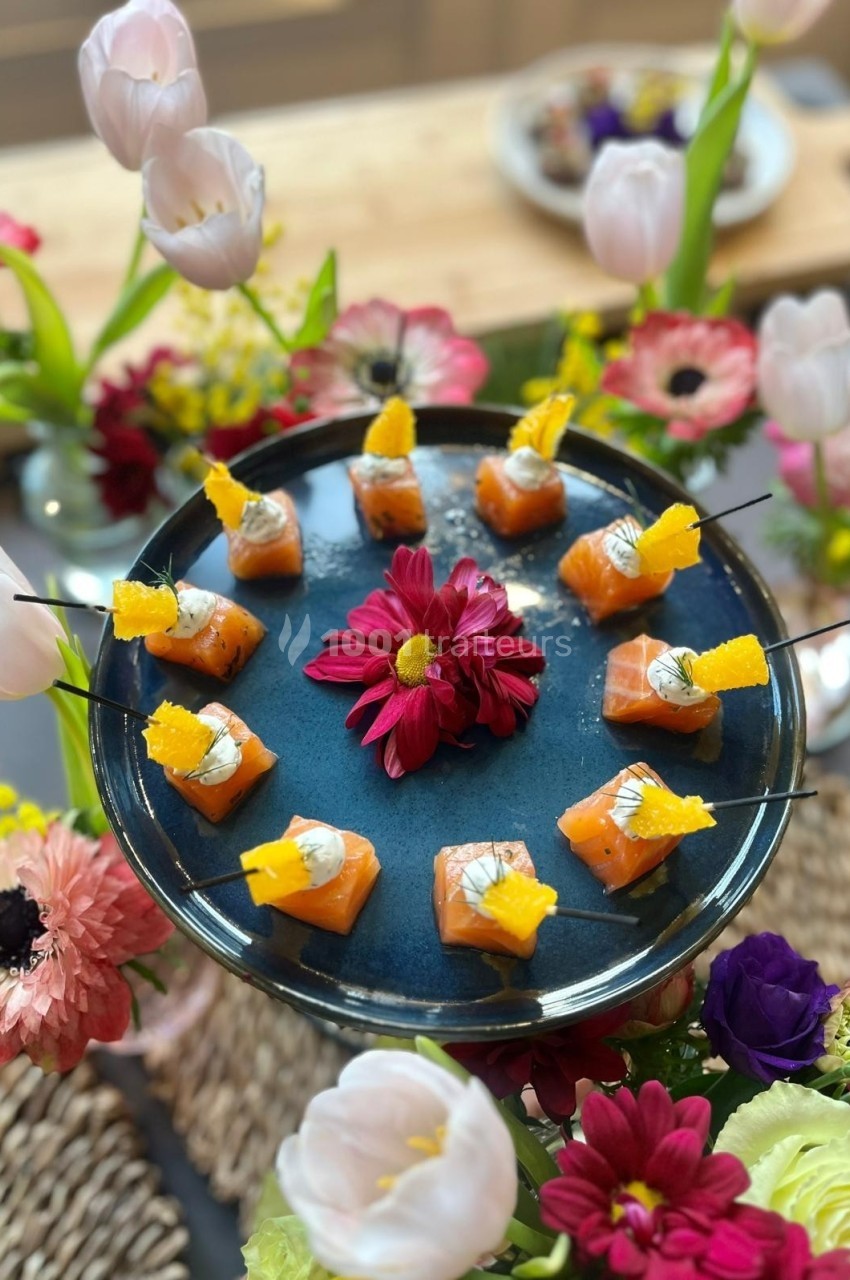 Assortiment de bouchées au saumon sur un plateau bleu, entouré de fleurs colorées en décoration.