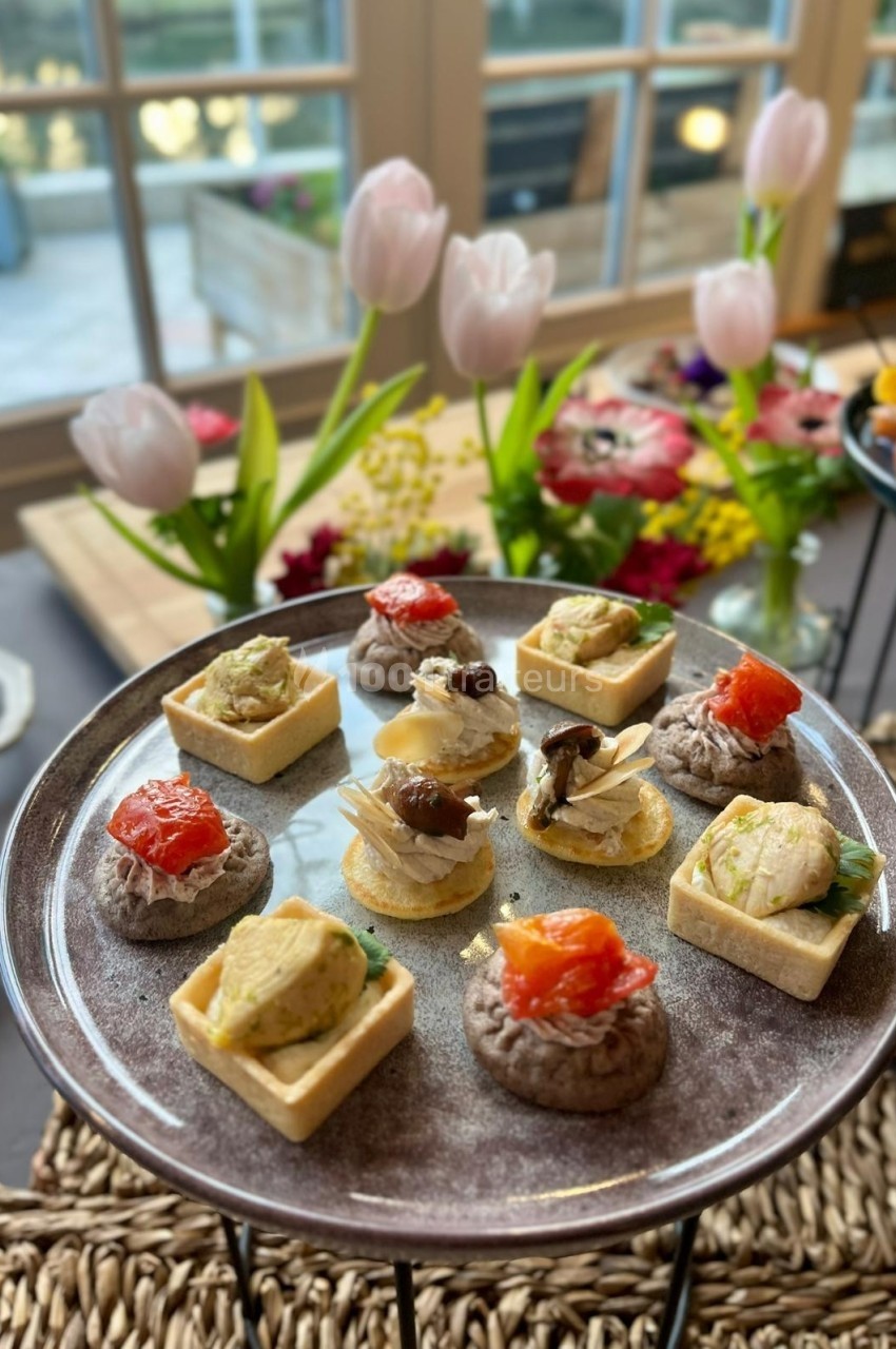 Plateau d'amuse-bouches variés sur une table décorée de fleurs, avec une vue sur un jardin en arrière-plan.