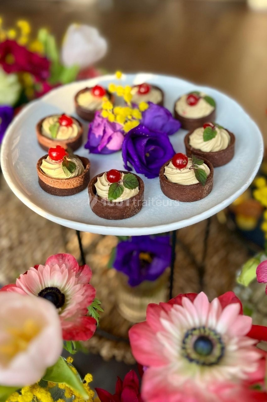 Assortiment de petits desserts chocolatés garnis de crème et de cerises, présentés sur un plat entouré de fleurs colorées.