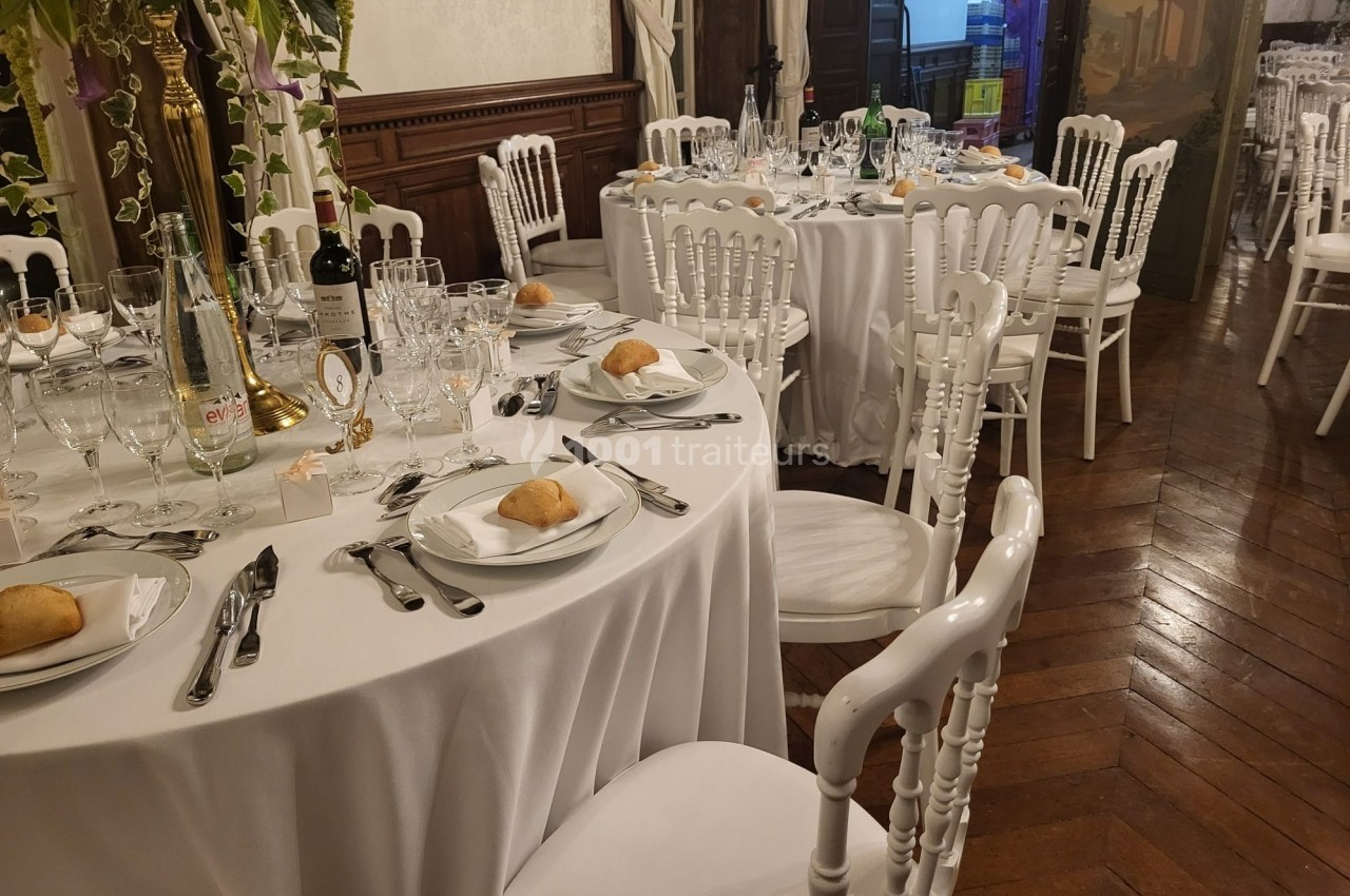 Salle de réception avec tables rondes dressées, nappes blanches, couverts, verres et chaises blanches sur parquet.