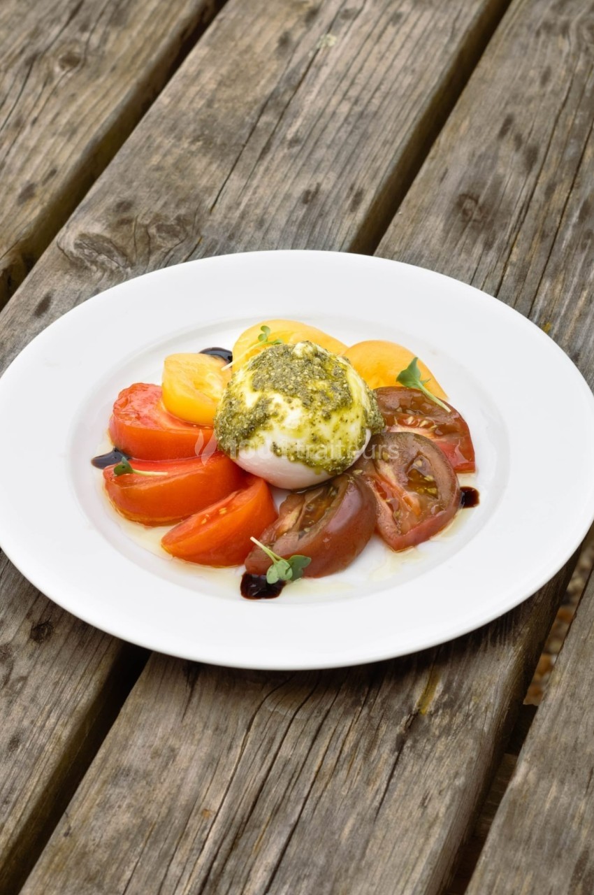 Assiette de burrata garnie de pesto, entourée de tranches de tomates rouges et jaunes sur une table en bois.