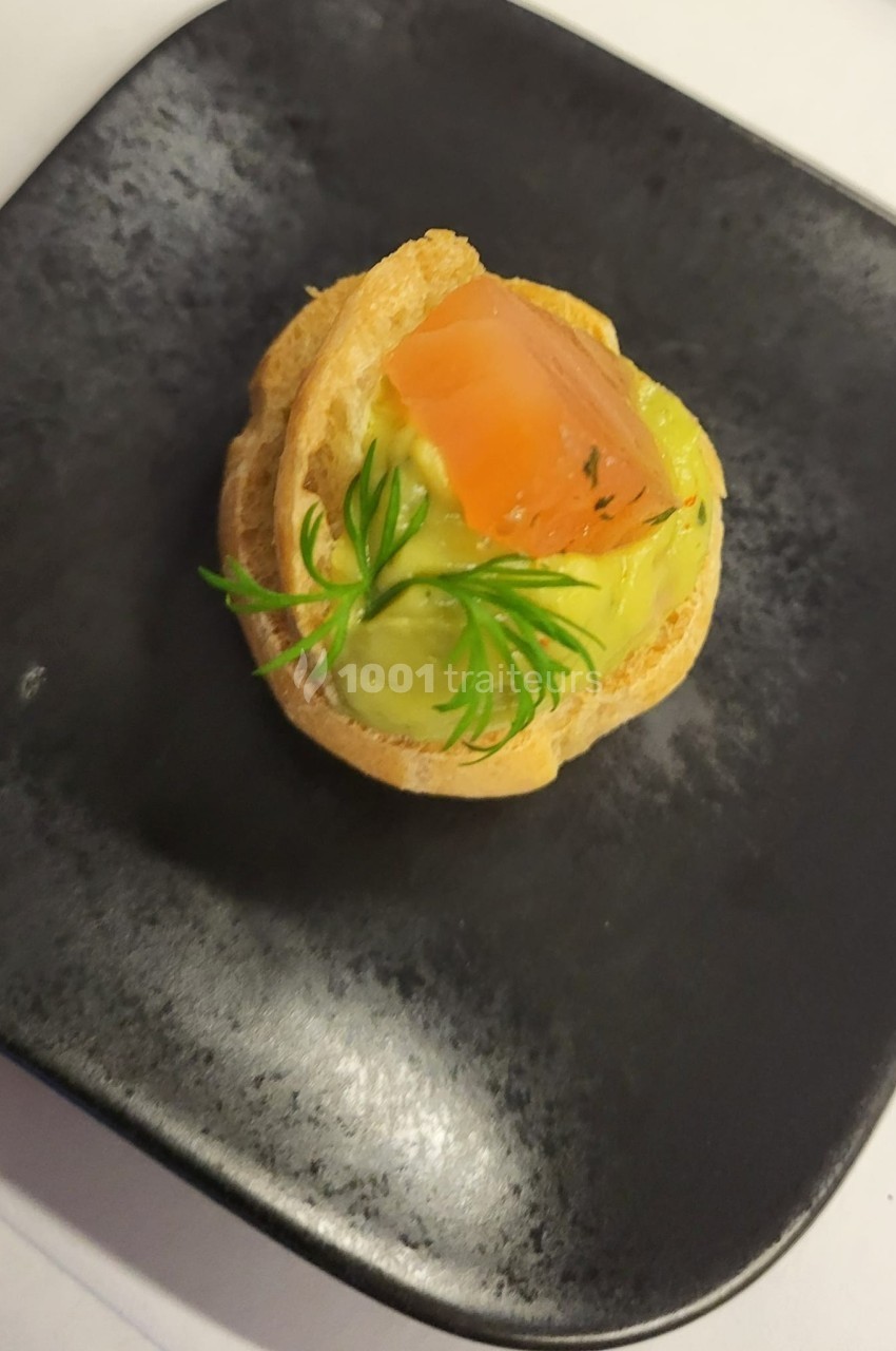 Chou garni de mousse verte, surmonté d'un morceau de saumon et décoré d'une herbe, présenté sur une assiette noire.