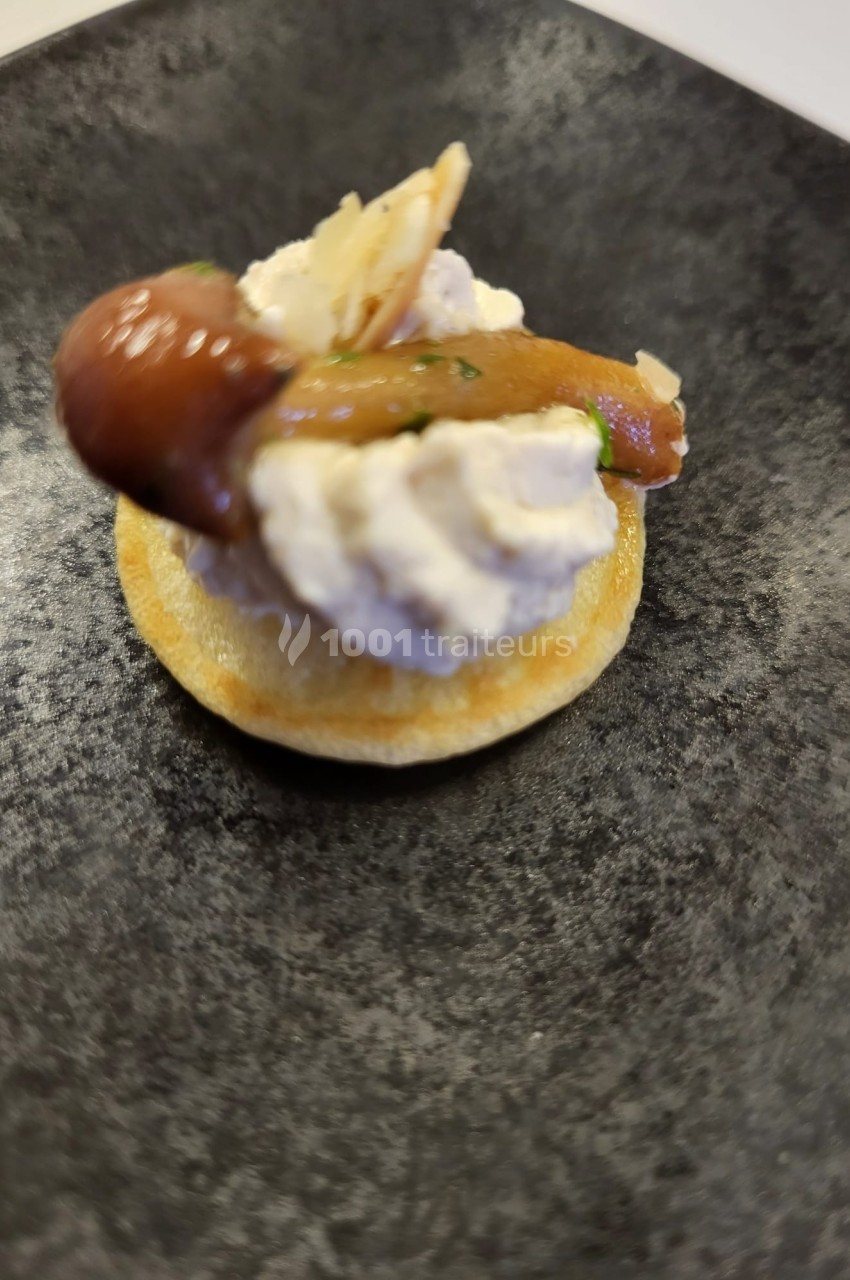 Blinis garni de fromage frais, champignons marinés et copeaux de fromage, présenté sur une assiette noire.