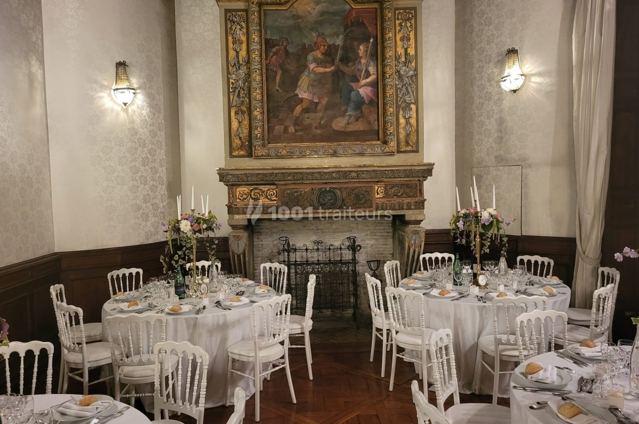 Salle élégante avec tables rondes dressées pour un repas, cheminée décorative et tableau ancien en arrière-plan.