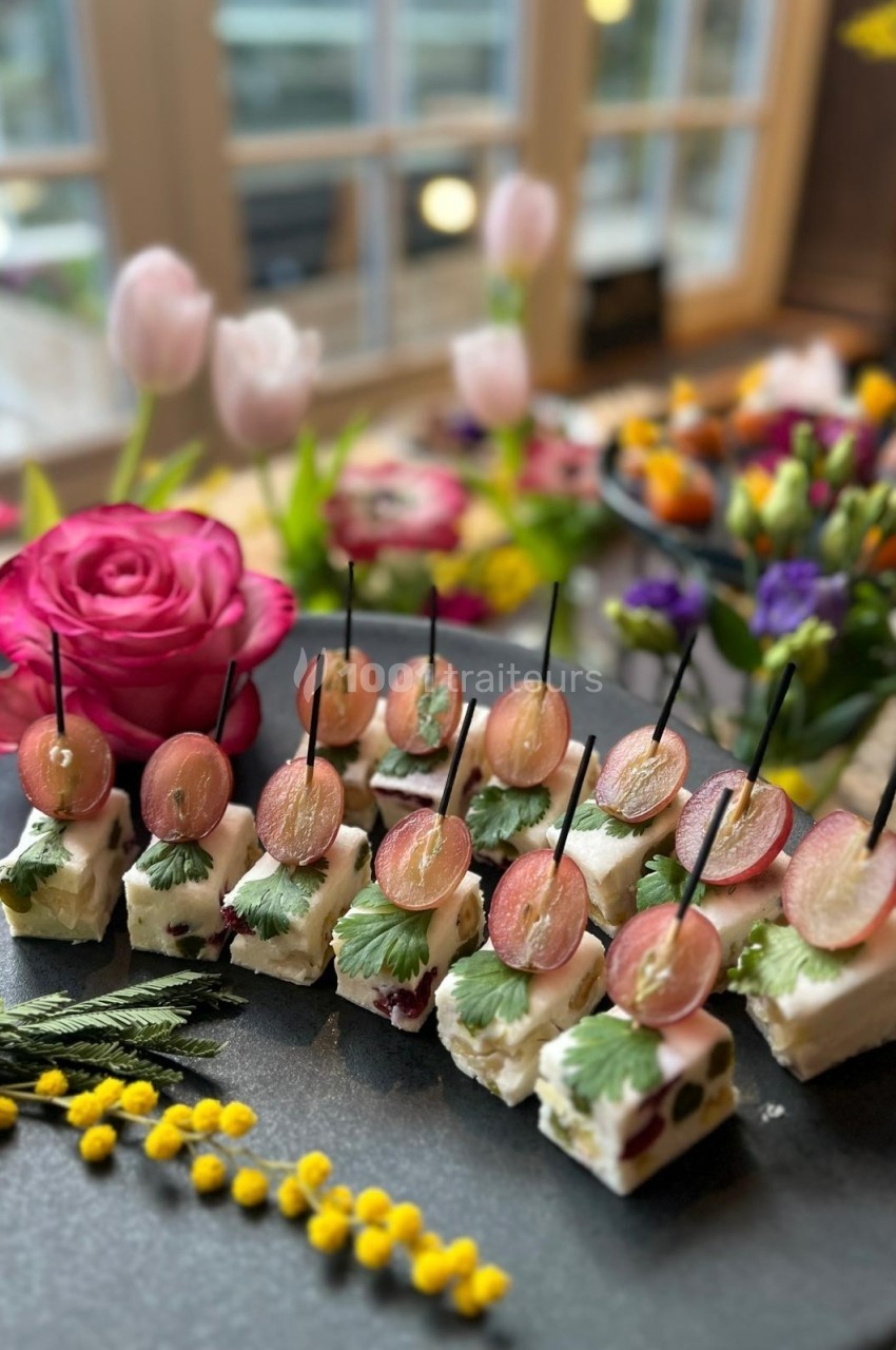 Assortiment de bouchées apéritives garnies de fromage, raisins et herbes, présenté sur un plateau avec décor floral.
