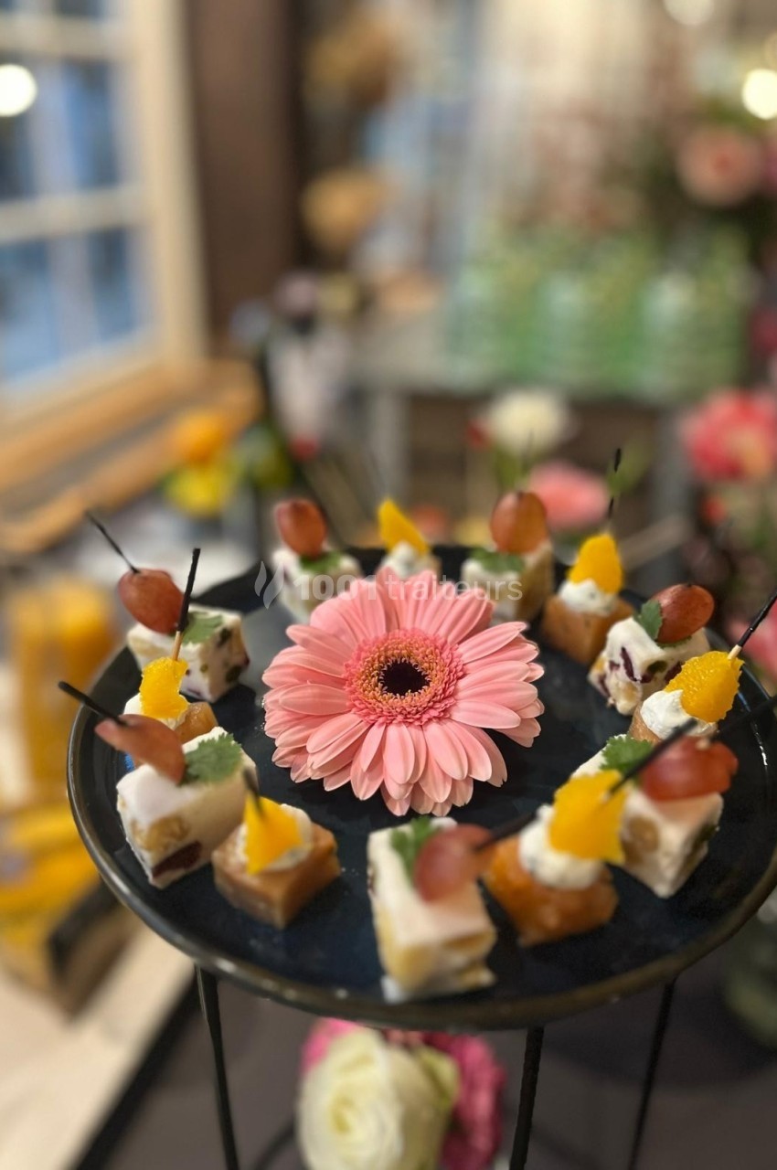 Plateau de bouchées apéritives décoré d'une fleur rose au centre, entouré de verrines en arrière-plan.