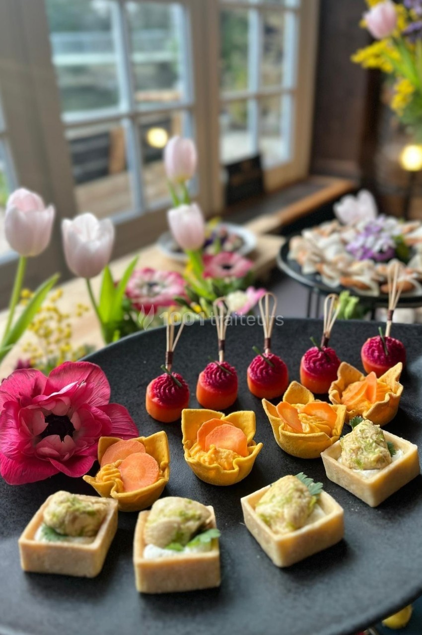 Plateau de bouchées apéritives variées présentées sur une table décorée de fleurs près d'une fenêtre.