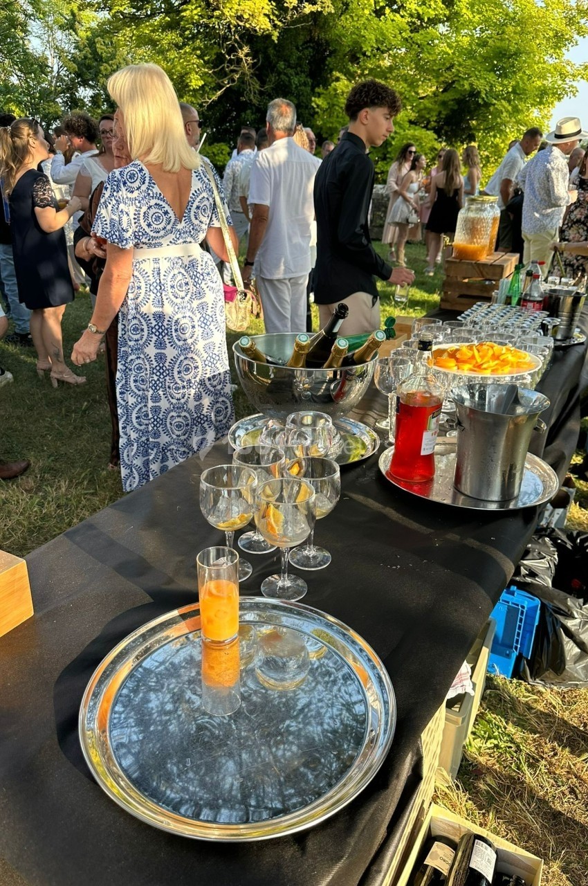Table de buffet en extérieur avec boissons, verres et carafes, entourée de convives dans un cadre verdoyant.