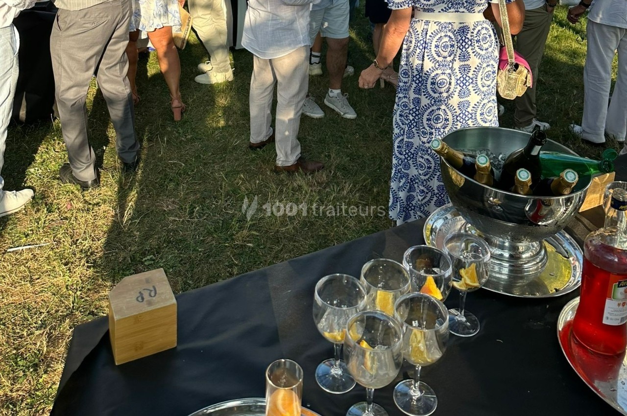 Personnes debout discutant dans un jardin, avec une table contenant des verres et un seau à champagne au premier plan.