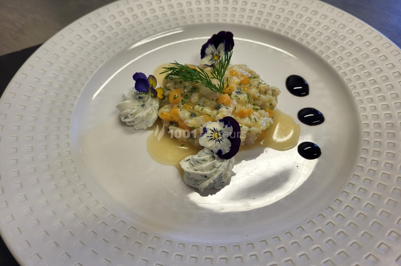 Tartare de poisson servi avec des fleurs comestibles, crème aux herbes et décorations de sauce sur une assiette blanche.