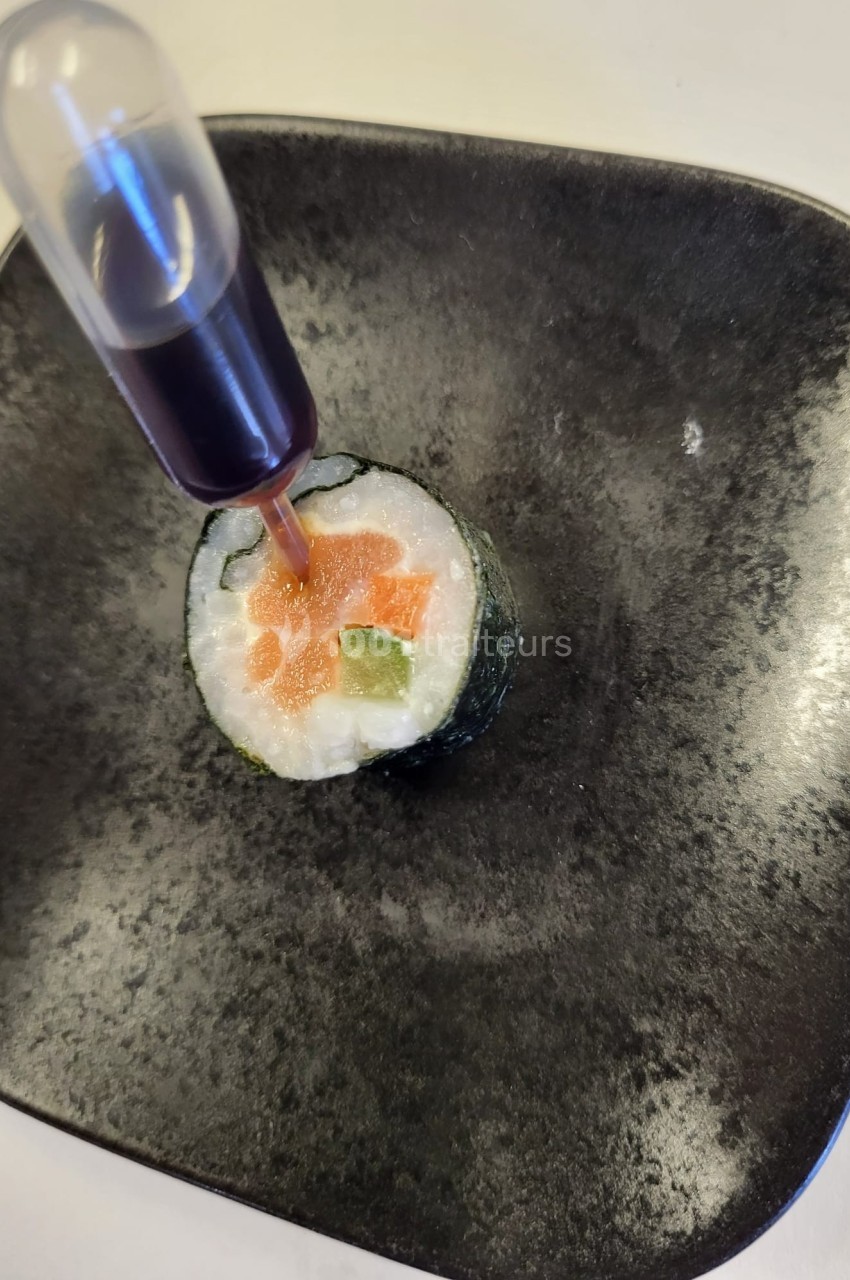 Sushi roulé avec saumon, avocat et riz, accompagné d'une pipette contenant une sauce foncée, présenté sur une assiette noire.