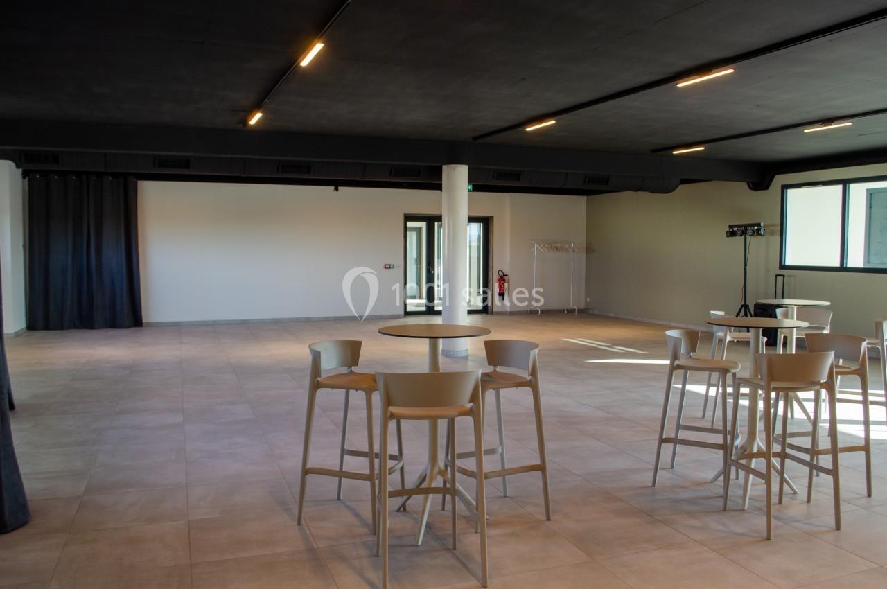 Salle spacieuse avec tables hautes, chaises et éclairage moderne, murs clairs et sol en carrelage beige.