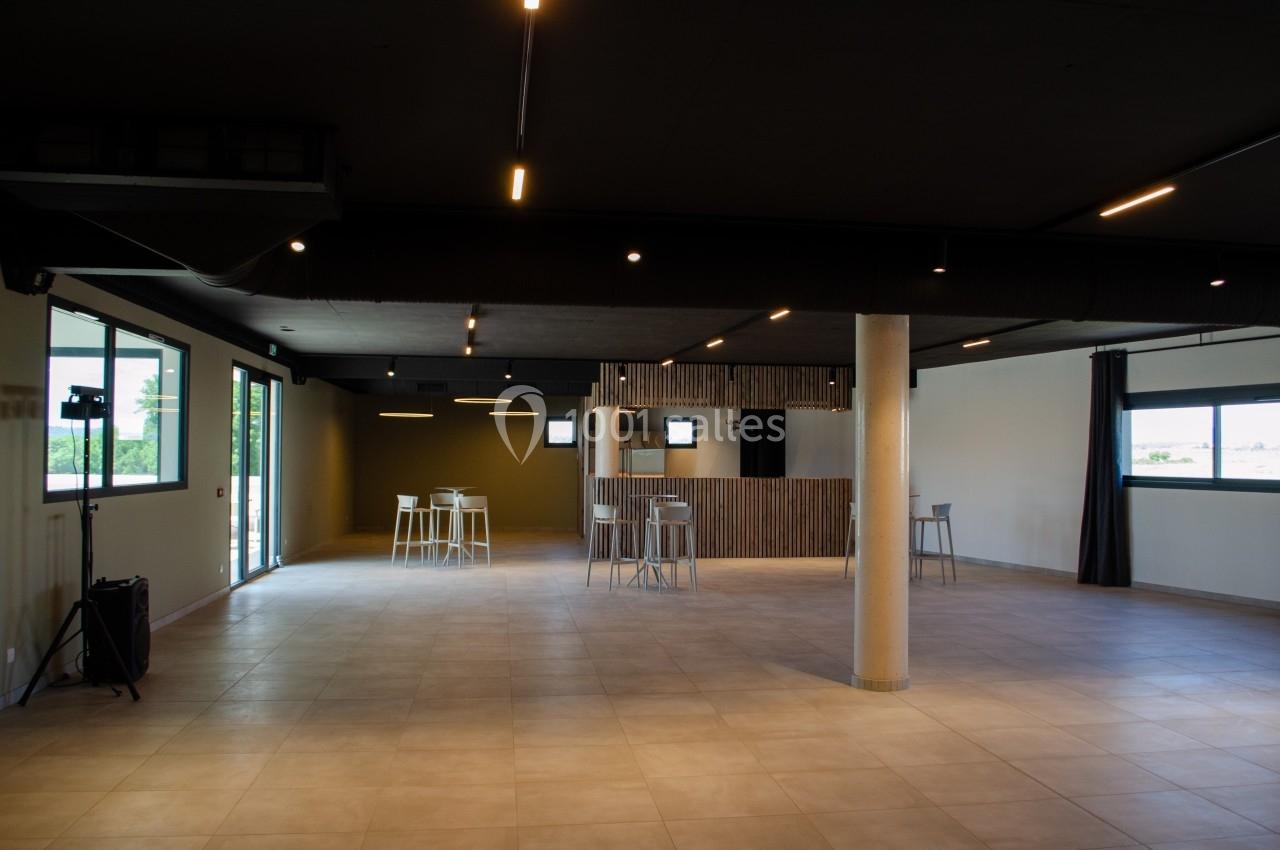 Salle spacieuse avec sol carrelé, éclairage moderne, grandes fenêtres et quelques tables hautes dispersées.