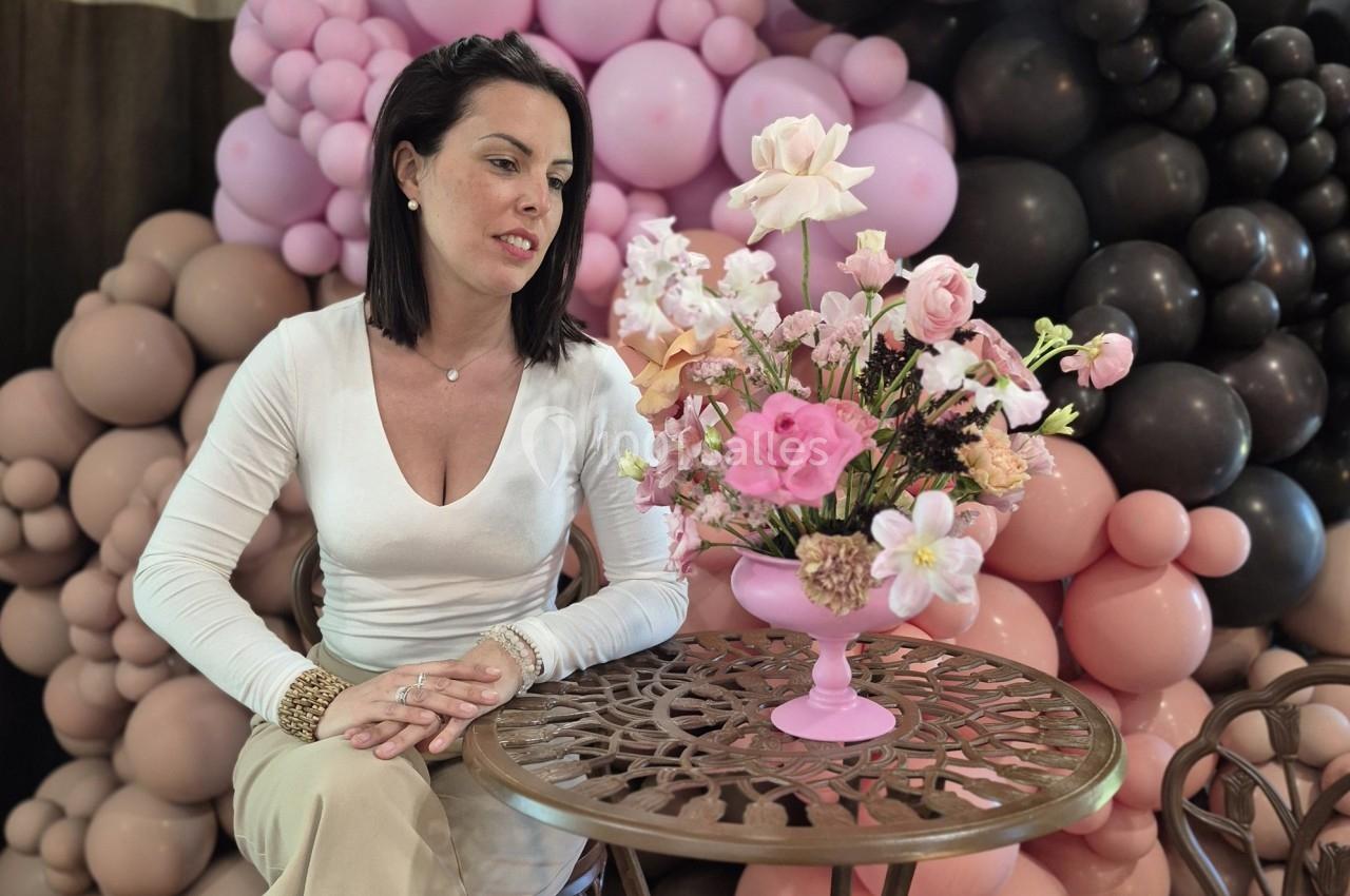 Une femme assise à une table regarde un bouquet de fleurs dans un vase rose, avec un décor de ballons en arrière-plan.