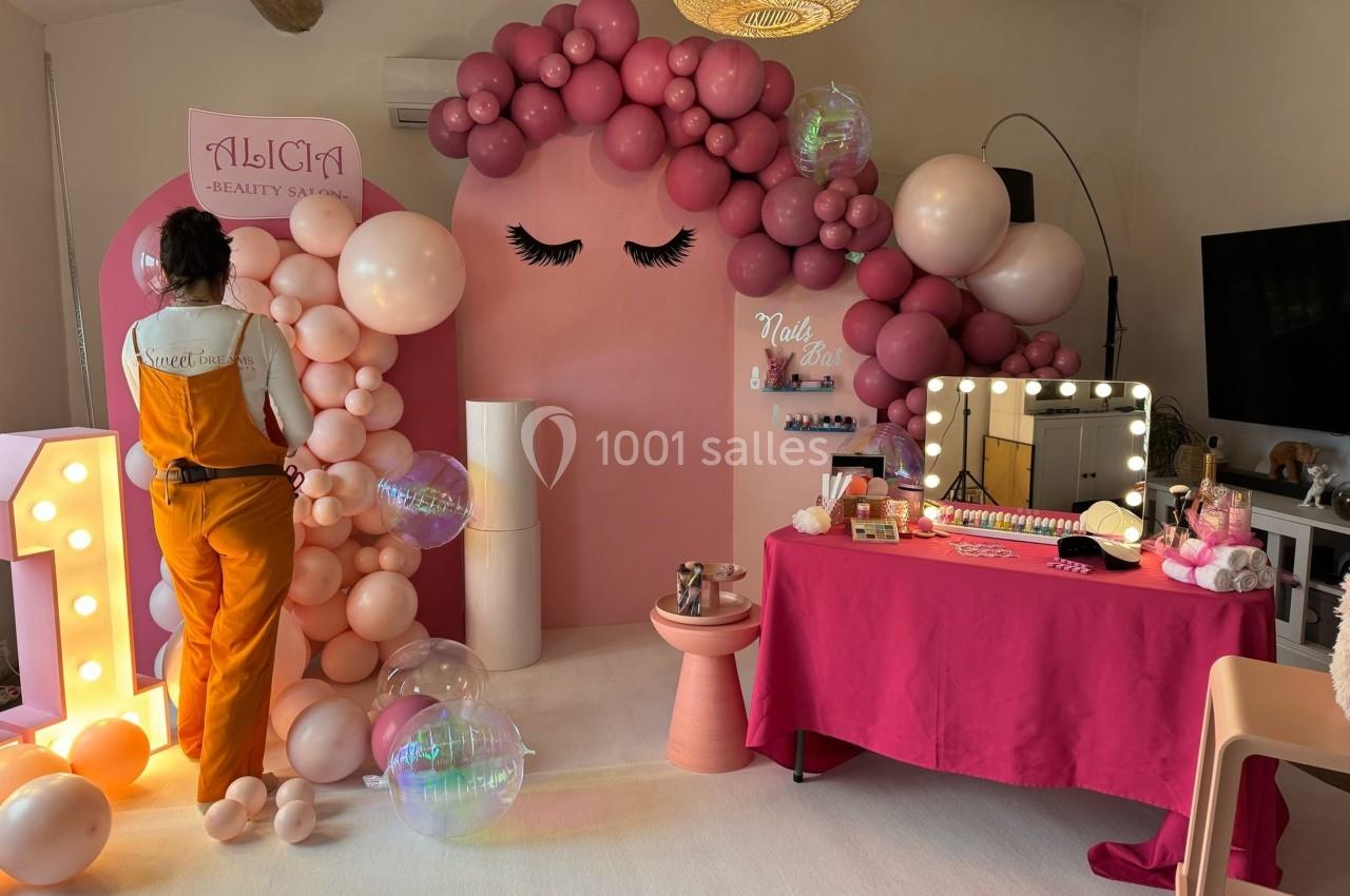 Décoration rose avec ballons, table de maquillage éclairée et personne en combinaison orange dans un salon esthétique.