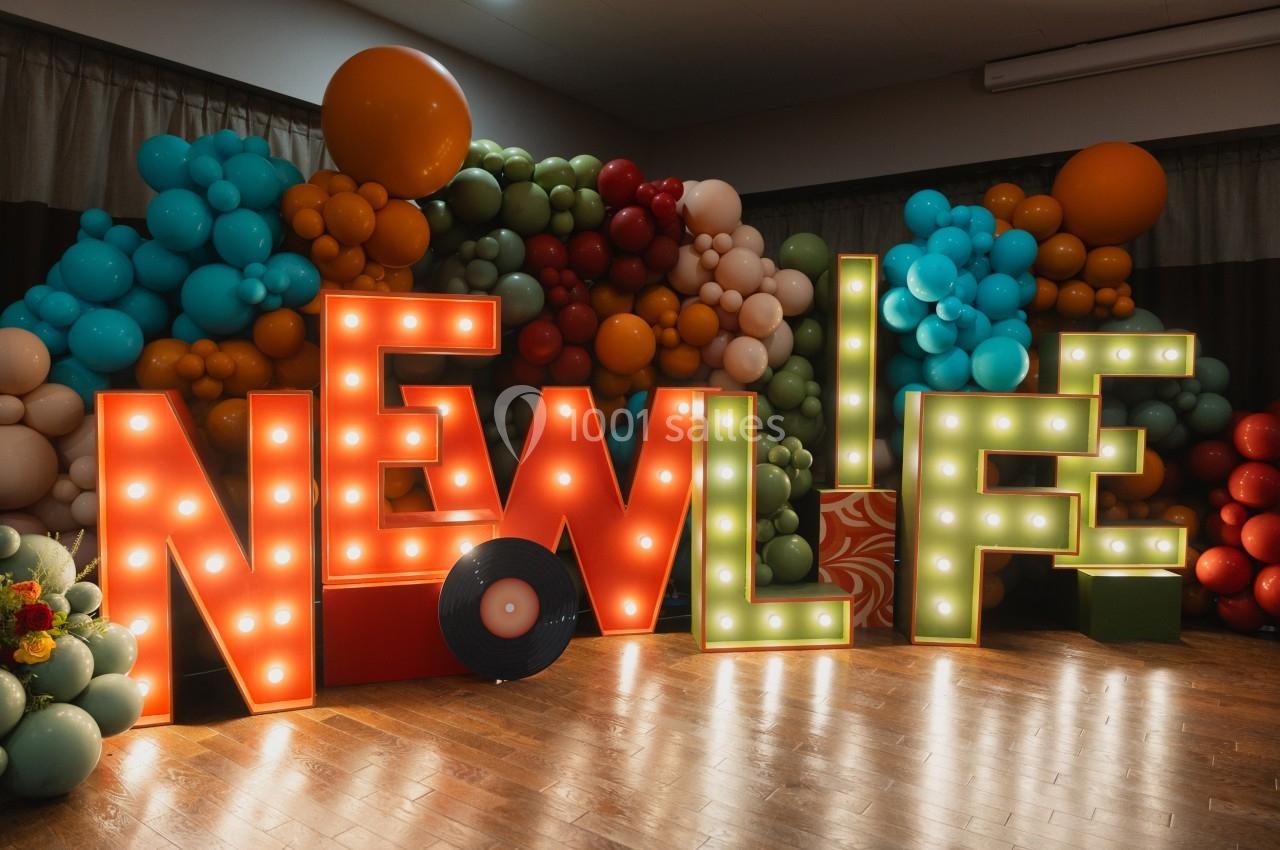 Lettres lumineuses formant ’NEW LIFE’ entourées de ballons colorés dans une salle décorée.