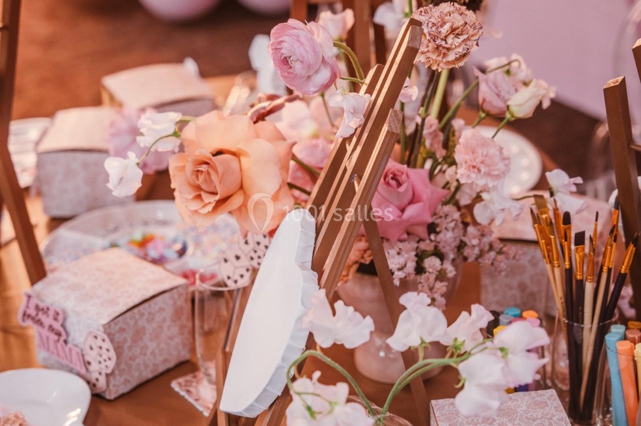 Atelier créatif avec chevalets, pinceaux, fleurs roses et matériel d'art disposés sur une table en bois.