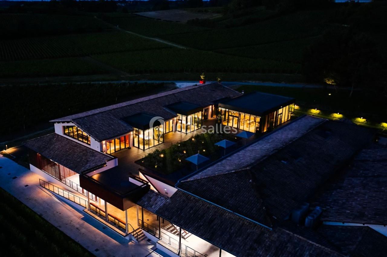 Vue aérienne nocturne d'un bâtiment moderne entouré de vignes, éclairé par des lumières extérieures.