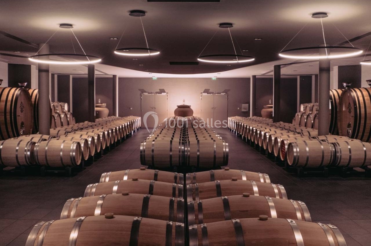 Rangées de fûts en bois alignés dans une cave à vin moderne, éclairée par des lumières suspendues.