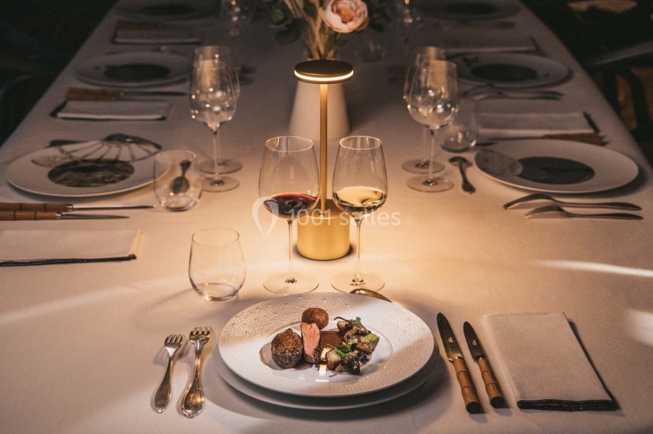 Table élégamment dressée avec assiettes, verres à vin et plat raffiné sous une lumière tamisée.