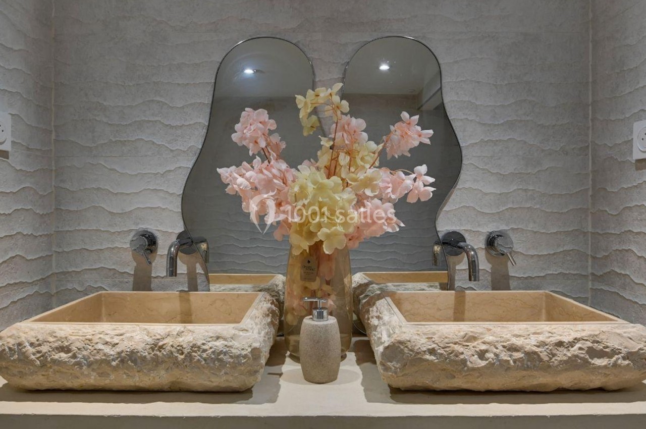 Deux lavabos en pierre avec un vase de fleurs roses et jaunes, placés devant un miroir ondulé.