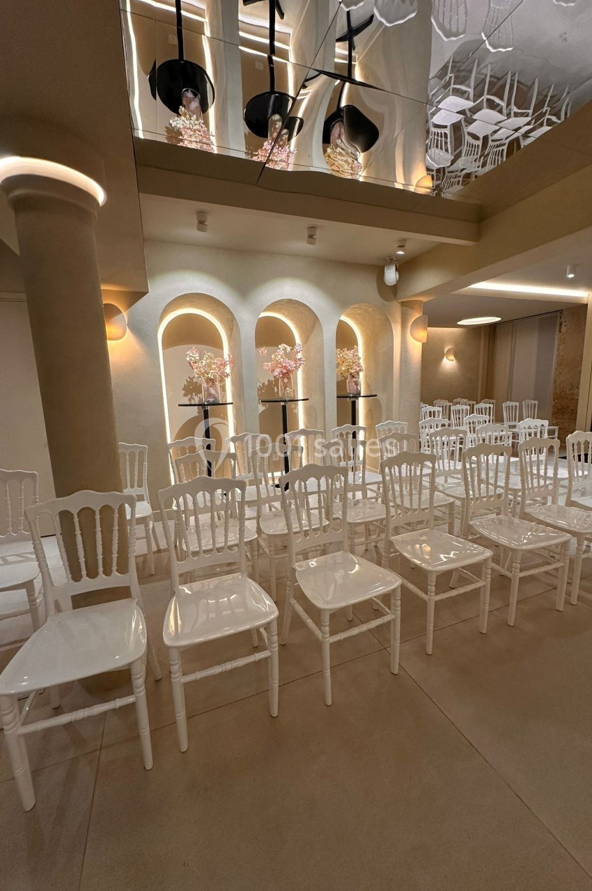 Salle décorée pour un événement avec des rangées de chaises blanches et des compositions florales sur des supports noirs.