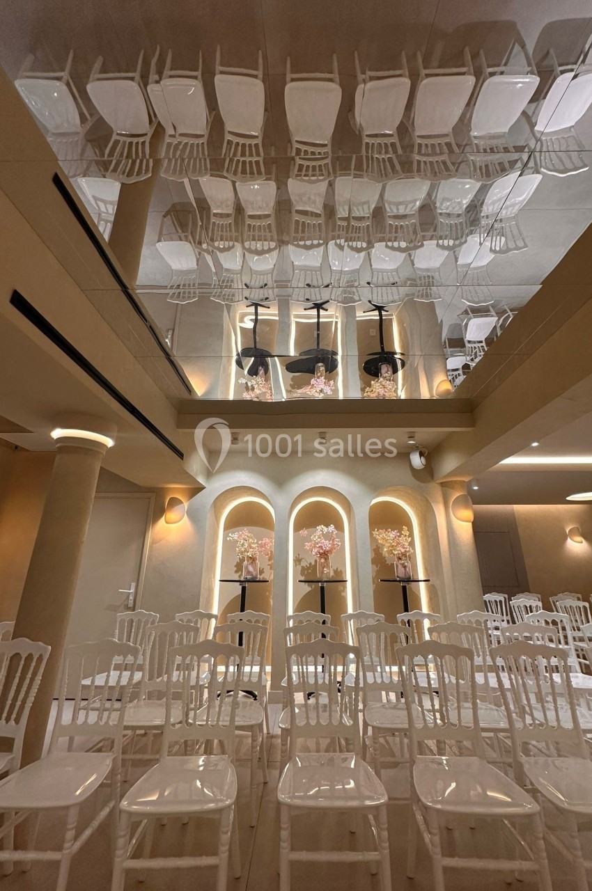 Salle de réception avec des rangées de chaises blanches et un plafond miroir reflétant l'ensemble de la décoration.