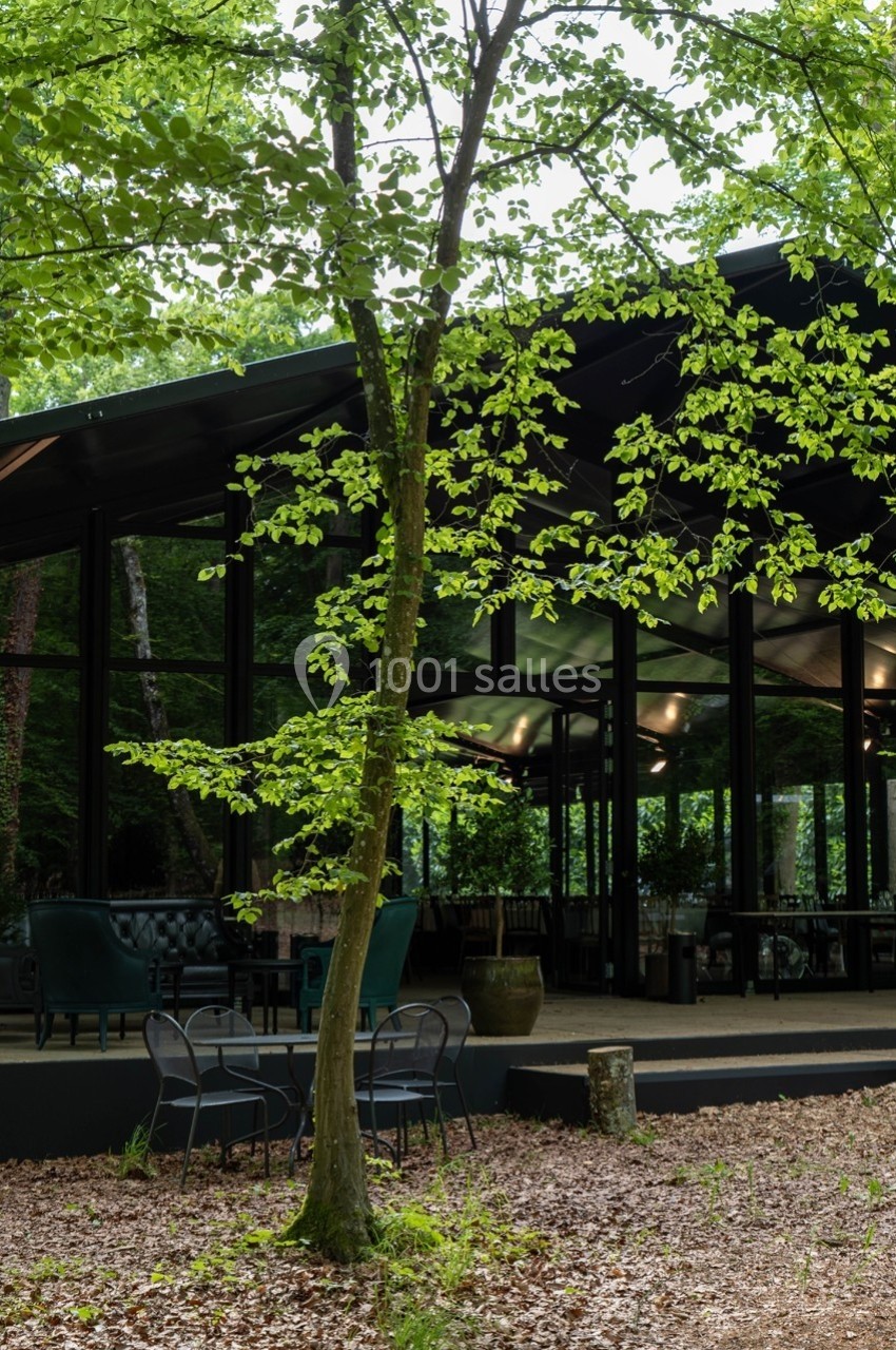 Façade vitrée d'un bâtiment moderne entouré de végétation, avec des chaises et une table en extérieur.