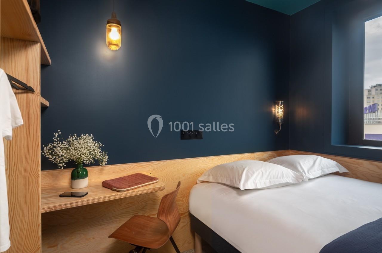 Chambre moderne avec un mur bleu, lit double, bureau en bois clair, chaise, lampe suspendue et fenêtre donnant sur l…